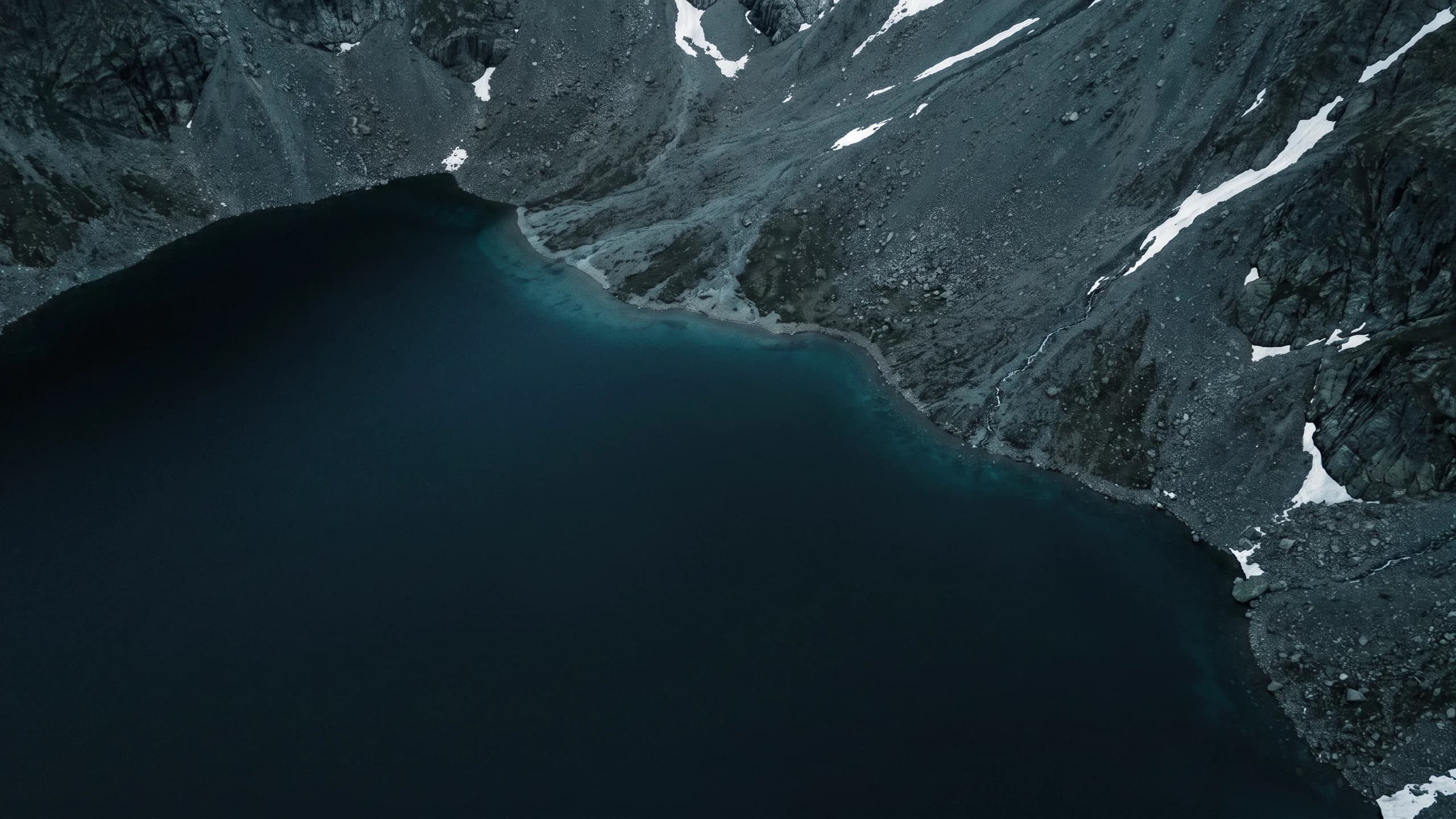 Aerial view of a dark alpine lake beside grey scree and patches of snow.